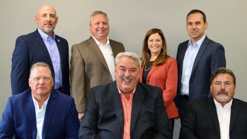 Pictured is the UIS Insurance & Investments management team. Four people in professional attire are standing behind three seated people in suits, all in front of a beige background.
